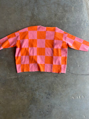 A pink and orange checkerboard long-sleeve top laid flat on a gray concrete floor