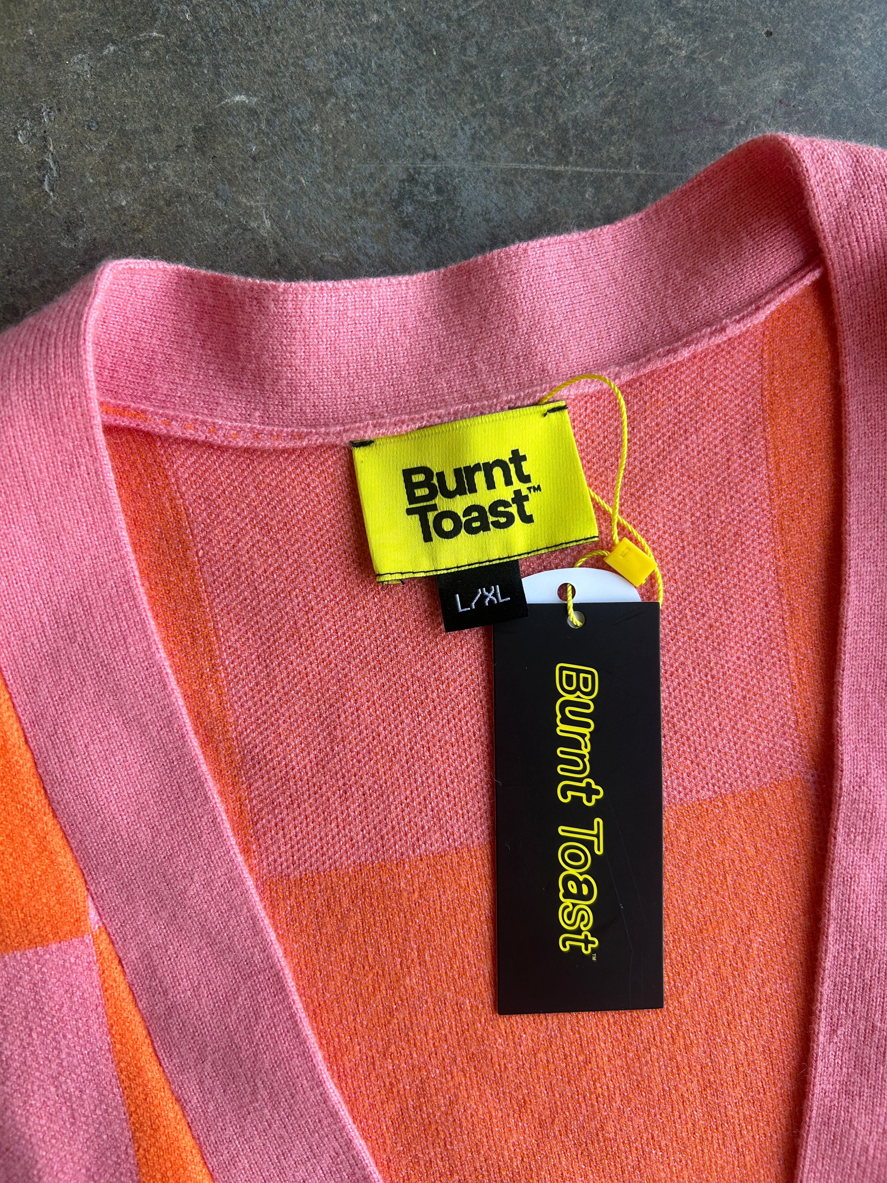 Close-up of a Burnt Toast knit sweater with a pink and orange checkered pattern, showing the yellow and black brand tags at the neckline