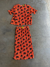 A two-piece matching set consisting of a short-sleeve top and a midi skirt, both in orange pleated fabric with a black animal print, laid flat on a gray concrete floor