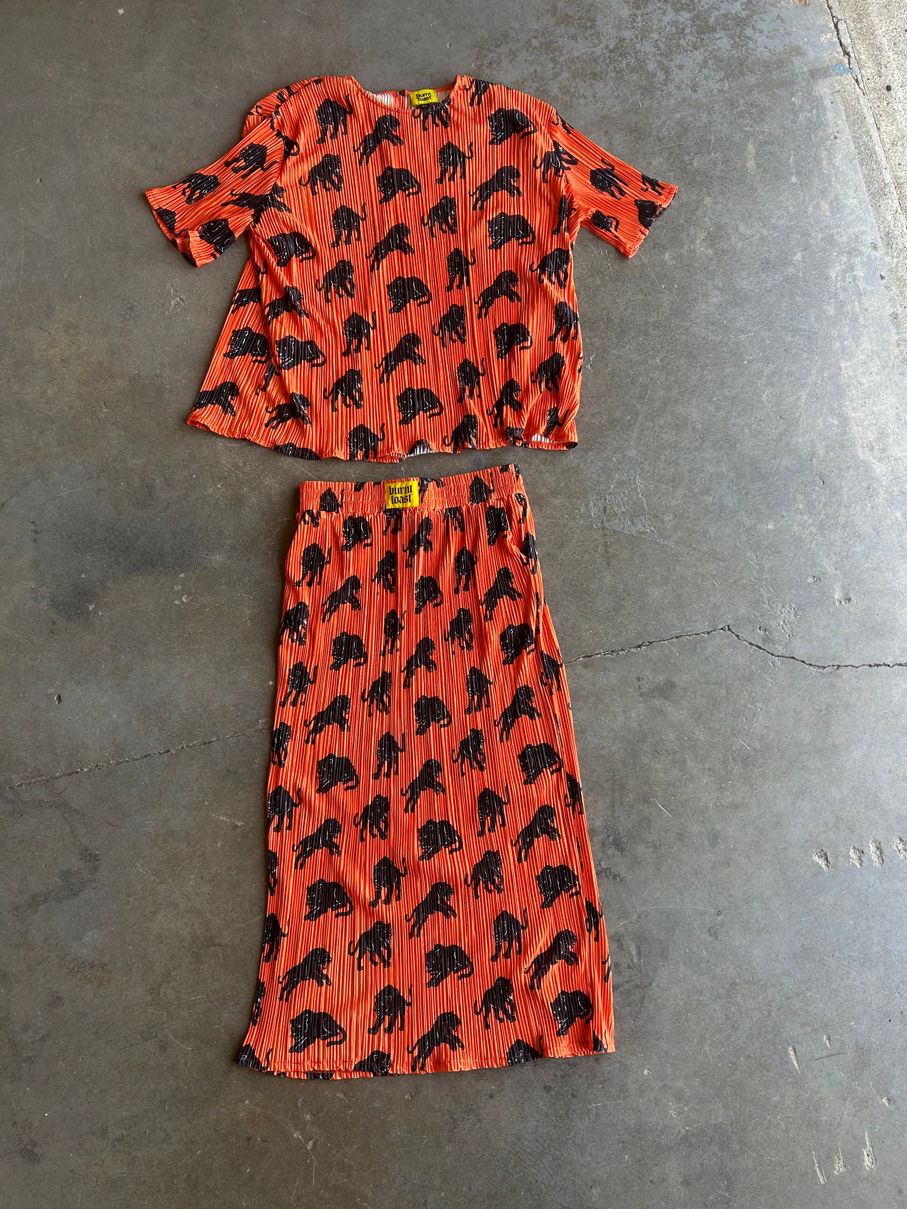A two-piece matching set consisting of a short-sleeve top and a midi skirt, both in orange pleated fabric with a black animal print, laid flat on a gray concrete floor