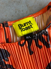 Close-up of a pleated orange garment with black abstract print and a bright yellow Burnt Toast brand label