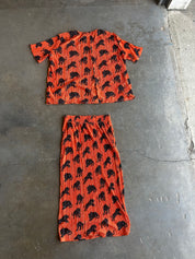 A two-piece matching set consisting of a short-sleeve button-up shirt and a pleated midi skirt, both in orange with a black animal print, laid flat on a gray concrete floor