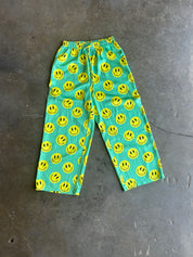 Bright turquoise blue pants with an all-over yellow smiley face pattern, laid flat on a gray concrete floor