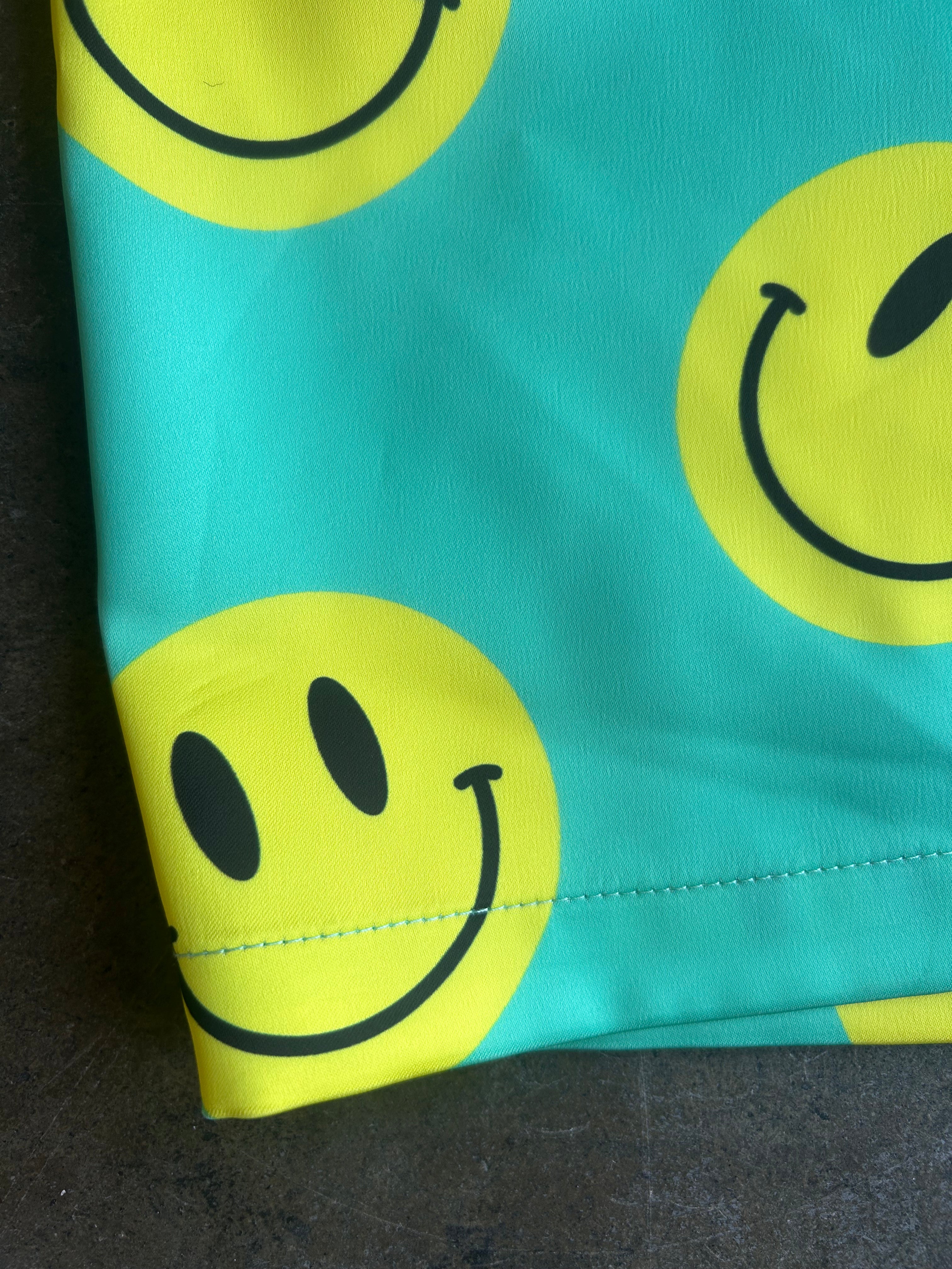 A close-up of a turquoise fabric with a repeating yellow smiley face pattern, showing a folded corner and stitching detail