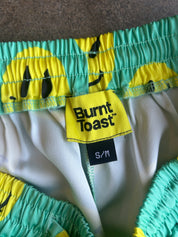Close-up of Burnt Toast swim trunks featuring a yellow and mint green sun pattern, with a yellow brand label and black size tag