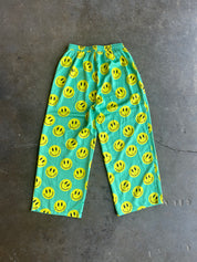 Bright turquoise blue pants covered in a repeating yellow smiley face pattern, laid flat on a dark gray concrete floor