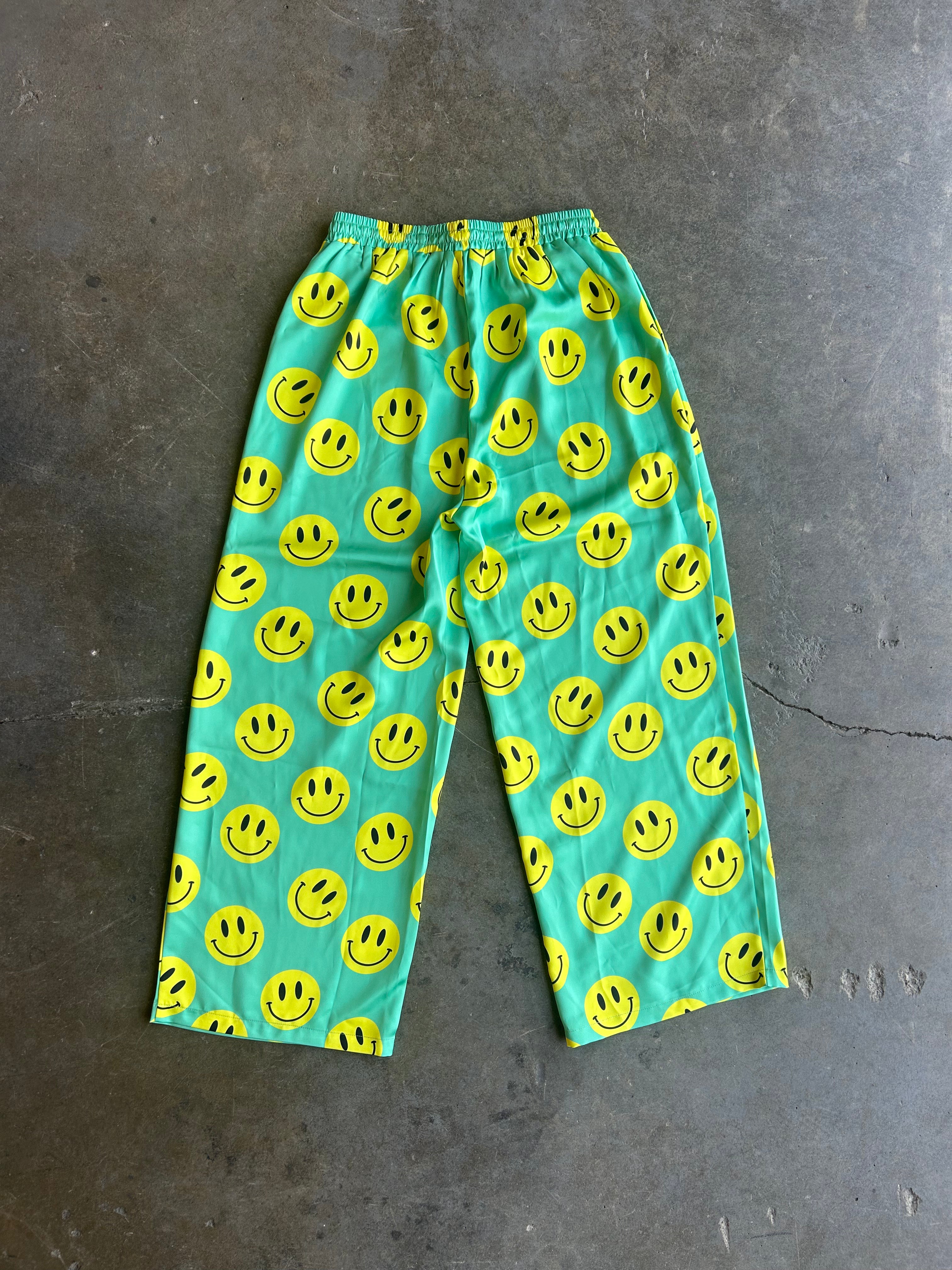 Bright turquoise blue pants covered in a repeating yellow smiley face pattern, laid flat on a dark gray concrete floor