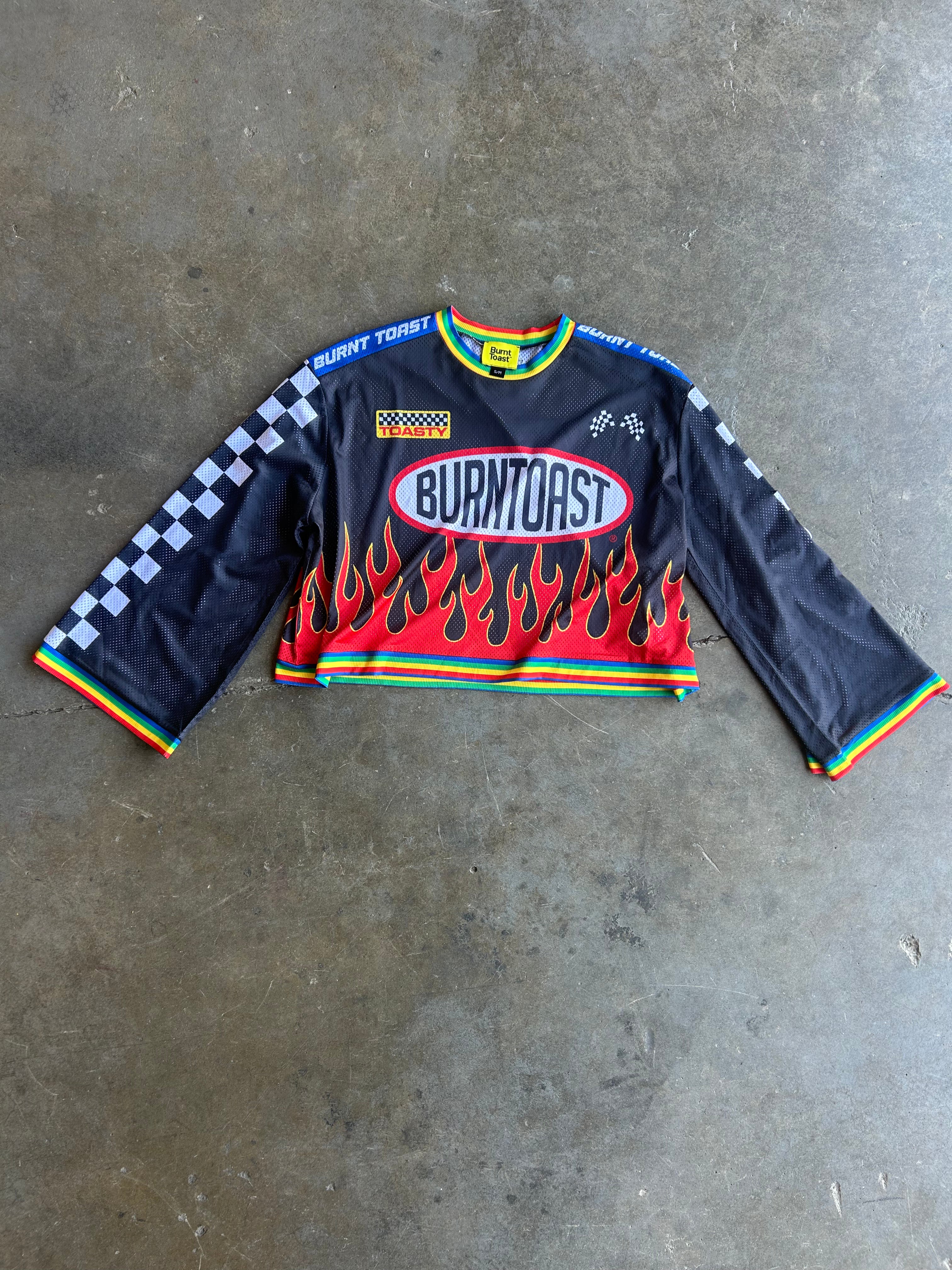 A long-sleeve black motocross jersey with 'Burntoast' branding, red and yellow flame graphics on the lower front, and a black-and-white checkered pattern on one sleeve, laid flat on a gray concrete floor