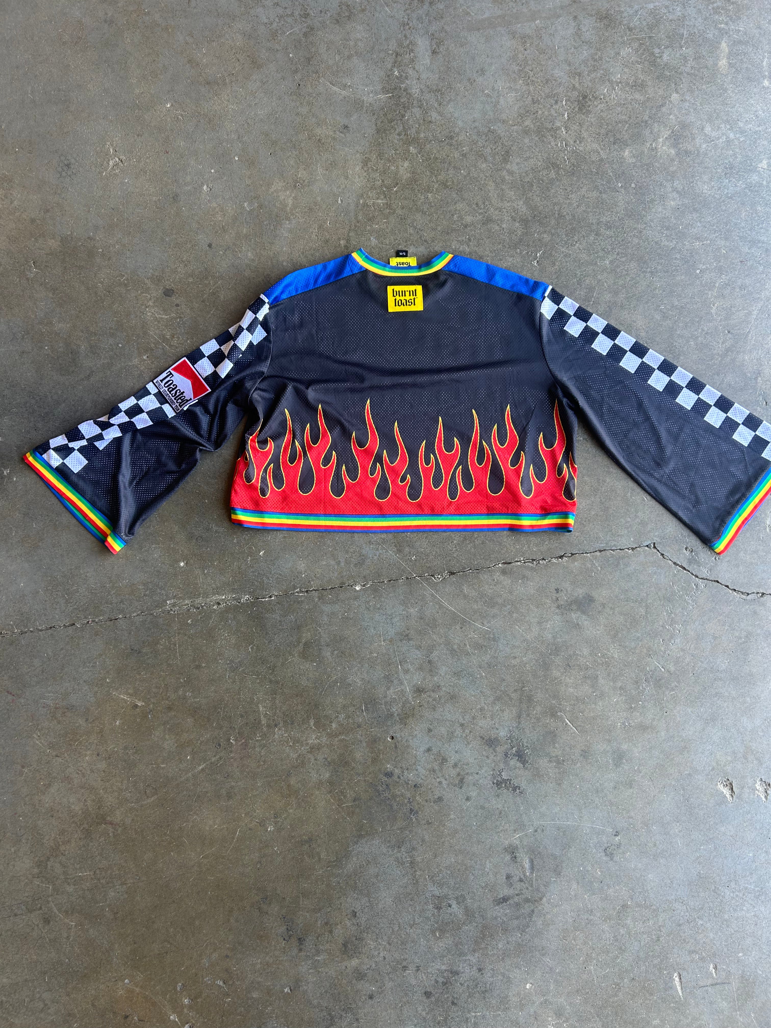 A long-sleeve motocross jersey laid flat on a concrete floor. The jersey is black with a red and orange flame pattern across the lower front, blue shoulder panels, a black-and-white checkered pattern on the sleeves, and a small yellow brand label at the collar