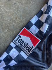 A close-up of a black and white checkered mesh jersey laid on a concrete surface, featuring a prominent embroidered patch that reads 'Toasted Wheel Championship Team' with a red and white logo