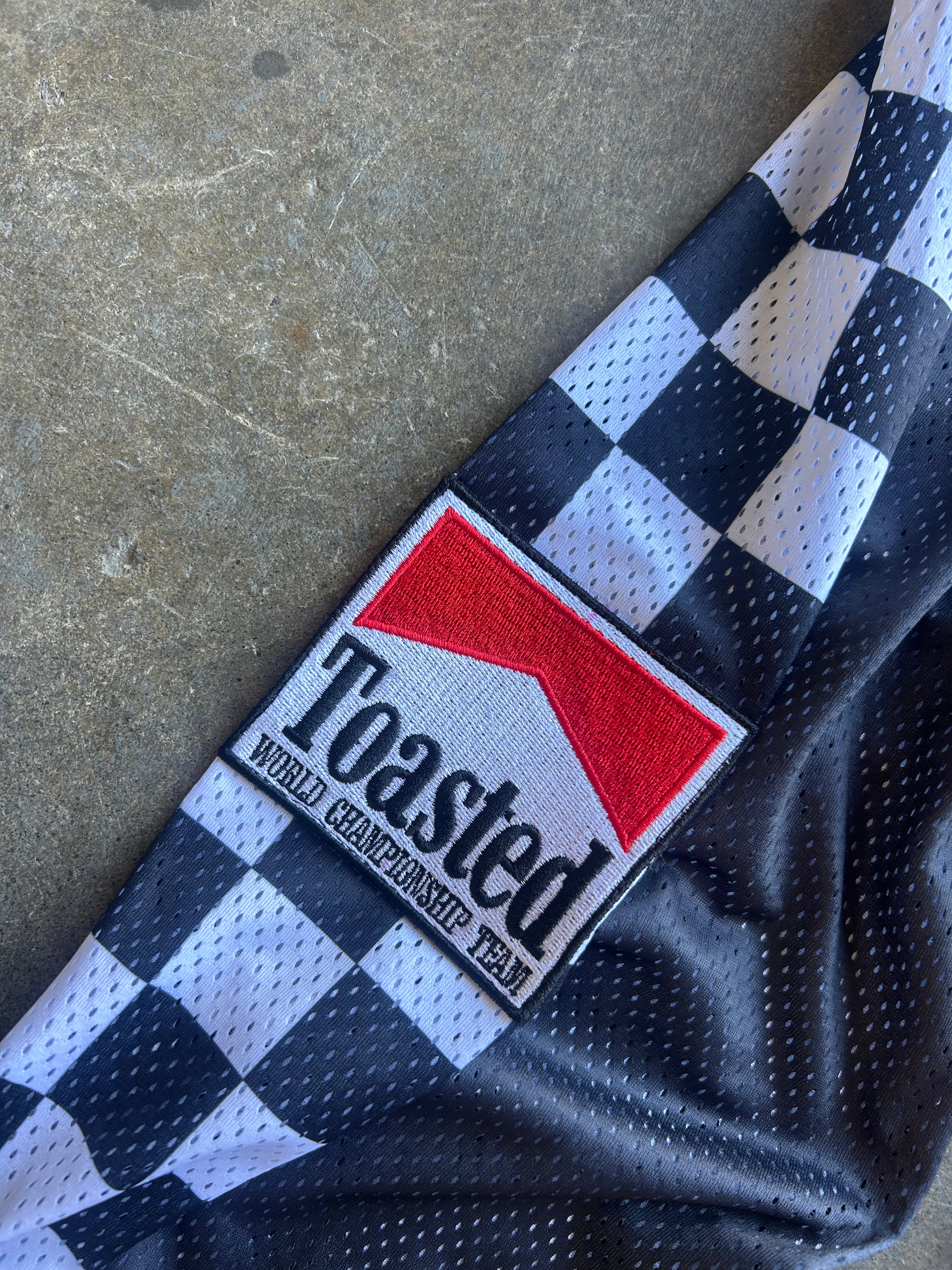 A close-up of a black and white checkered mesh jersey laid on a concrete surface, featuring a prominent embroidered patch that reads 'Toasted Wheel Championship Team' with a red and white logo