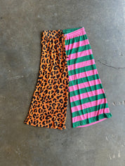 A pair of wide-leg cropped pants laid flat on a gray concrete floor, featuring a split design with an orange leopard print panel on the left leg and pink and green horizontal stripes on the right leg