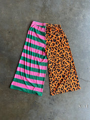 A pair of wide-leg cropped pants laid flat on a gray concrete floor, featuring a split design with pink and green horizontal stripes on one leg and an orange leopard print on the other