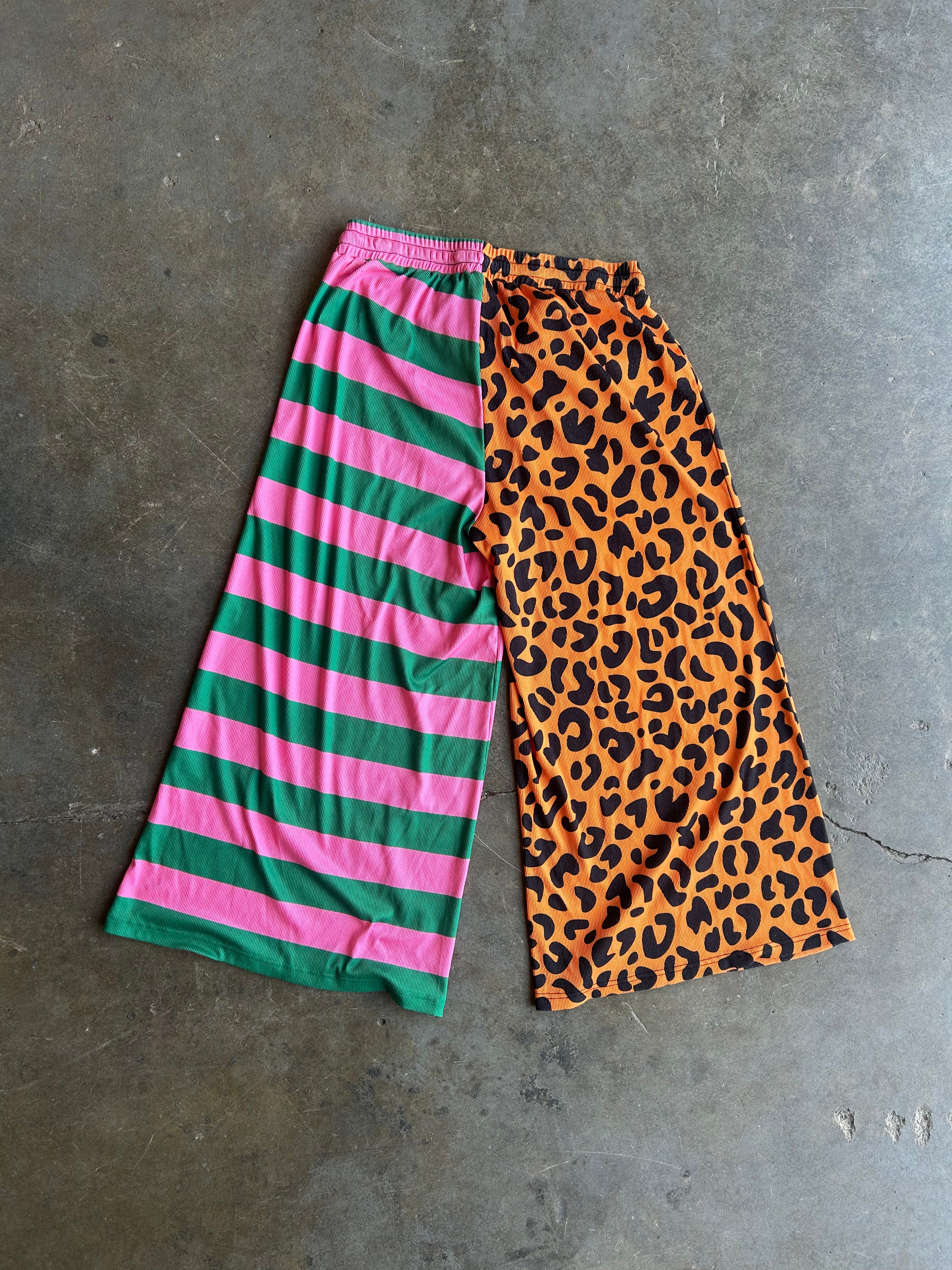 A pair of wide-leg cropped pants laid flat on a gray concrete floor, featuring a split design with pink and green horizontal stripes on one leg and an orange leopard print on the other