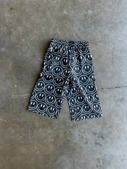 Black and white patterned shorts with a repeating smiley face design, laid flat on a textured concrete floor