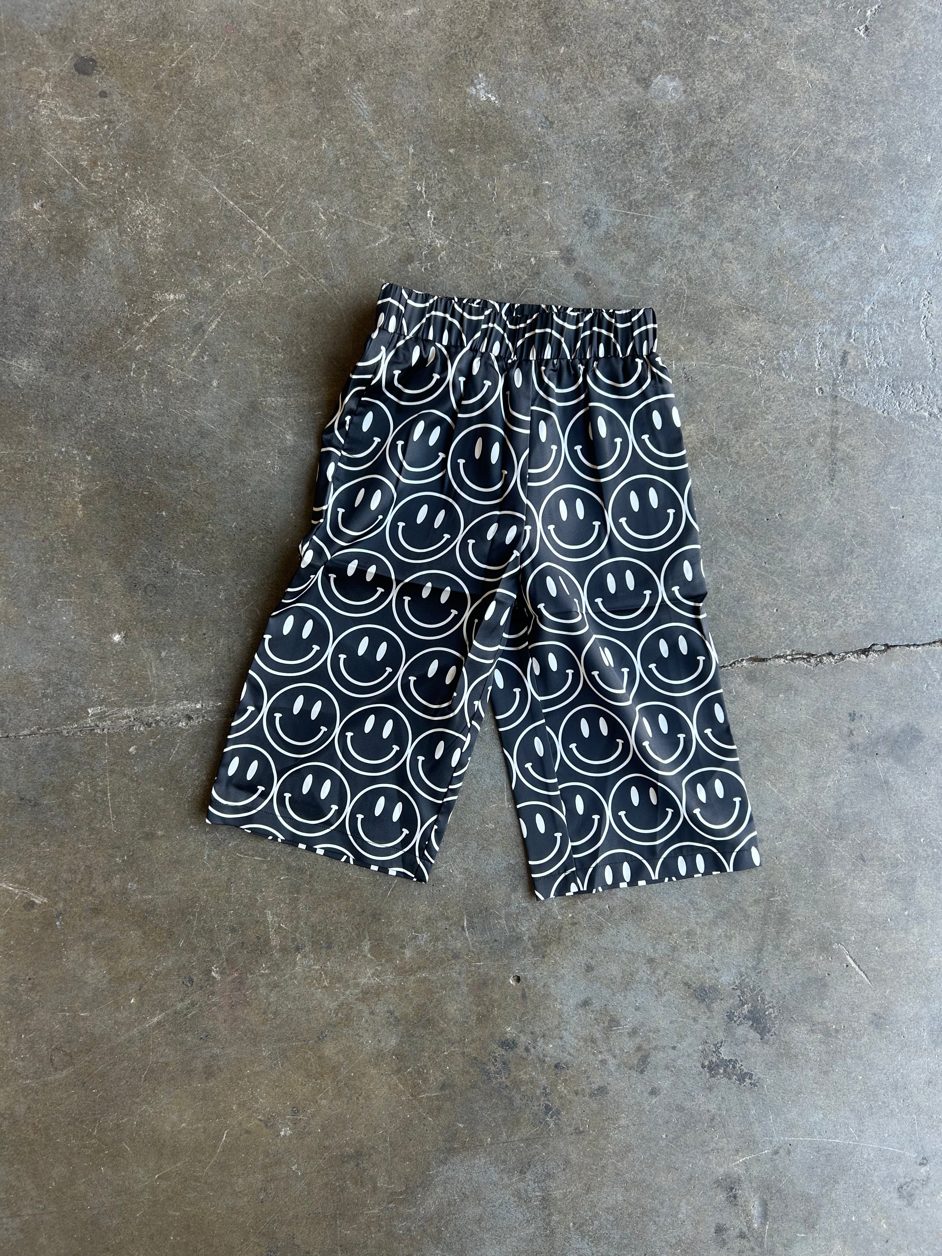 Black and white patterned shorts with a repeating smiley face design, laid flat on a textured concrete floor