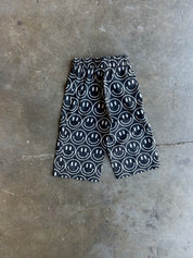 Black and white patterned shorts with a repeating smiley face design, laid flat on a gray concrete floor