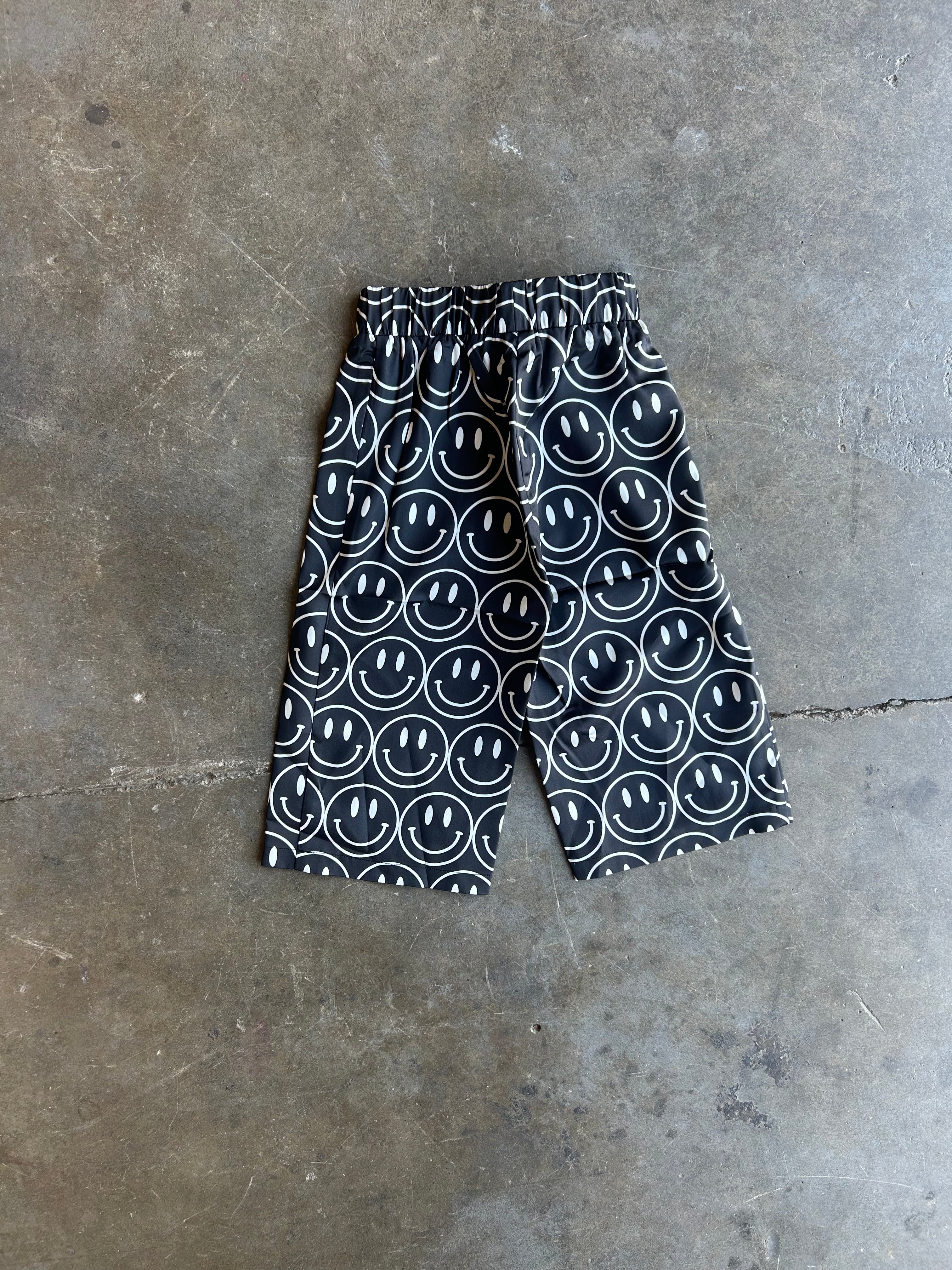 Black and white patterned shorts with a repeating smiley face design, laid flat on a gray concrete floor