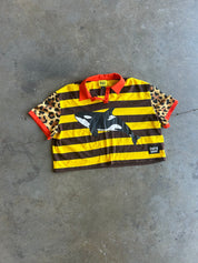 A short-sleeve striped shirt laid flat on a gray concrete floor. The shirt has bold yellow and dark brown horizontal stripes, leopard-print sleeves, and red collar/cuff trim and a killer whale on the front