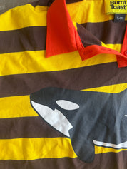A close-up of a Burnt Toast polo shirt featuring bold yellow and dark brown horizontal stripes, a bright red collar, and a large black and white orca graphic printed across the front