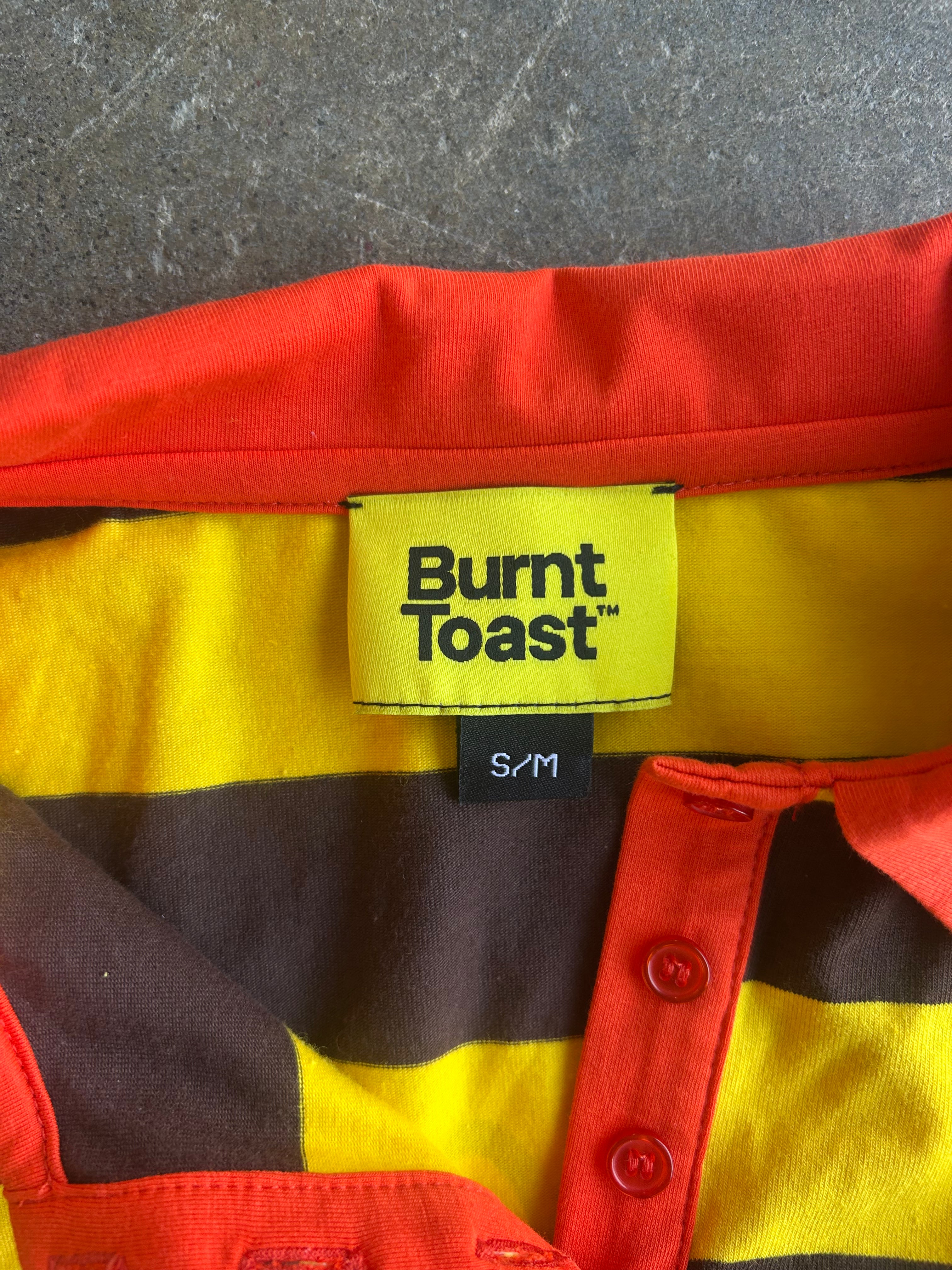 Close-up of a Burnt Toast striped shirt with yellow, brown, and red horizontal stripes, featuring a yellow brand label and red buttons
