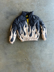 A black and beige flame-patterned puffer jacket with a yellow zipper, laid flat on a gray concrete floor
