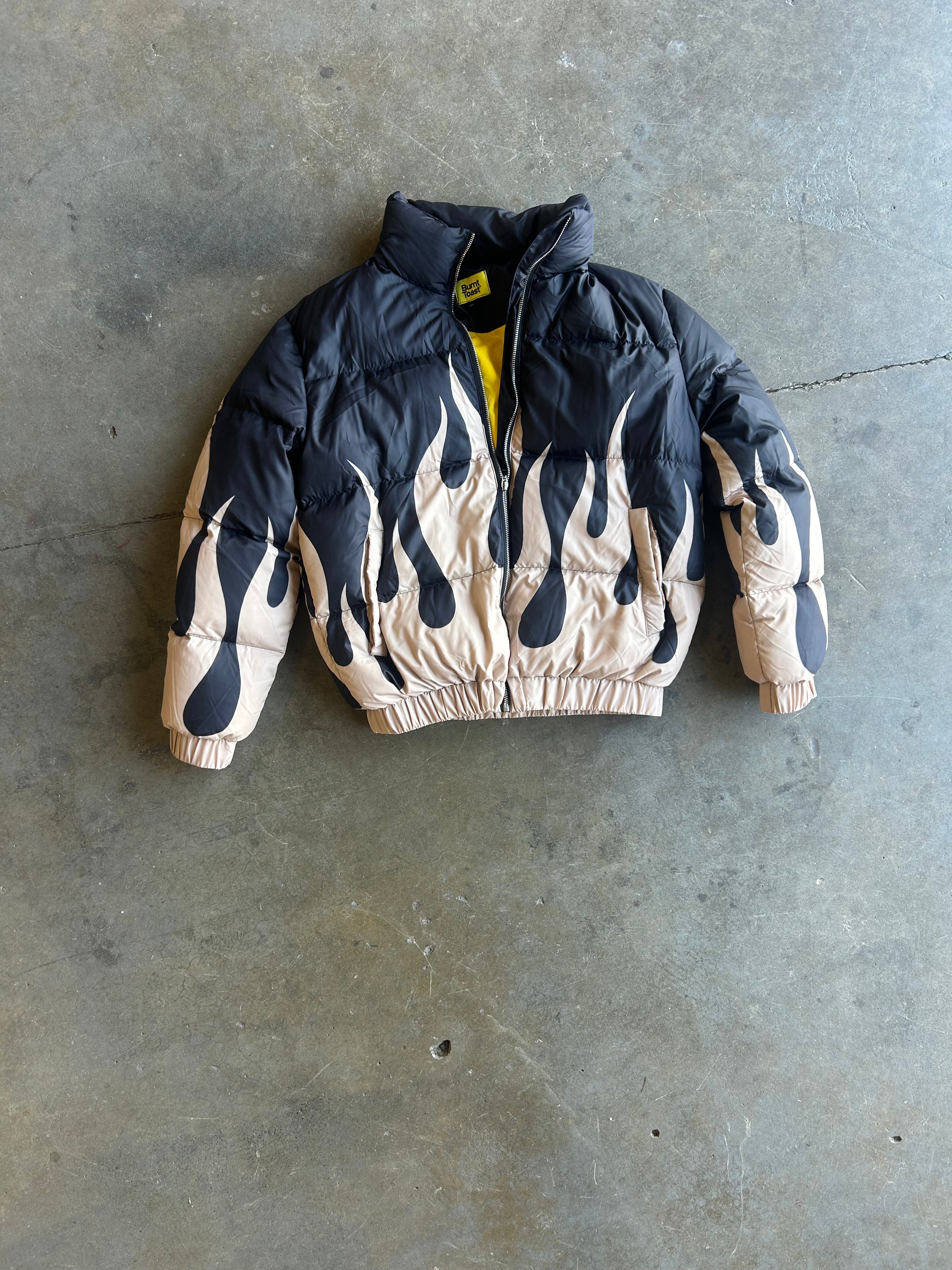 A black and beige flame-patterned puffer jacket with a yellow zipper, laid flat on a gray concrete floor