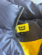 Close-up of a puffer jacket showing the yellow Burnt Toast brand label stitched onto the dark gray exterior next to a silver zipper, with a bright yellow panel visible at the bottom