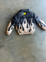 A puffer jacket laid flat on a concrete floor, featuring a black upper body and sleeves with a beige flame pattern on the lower body and cuffs. A small yellow label is visible at the back collar