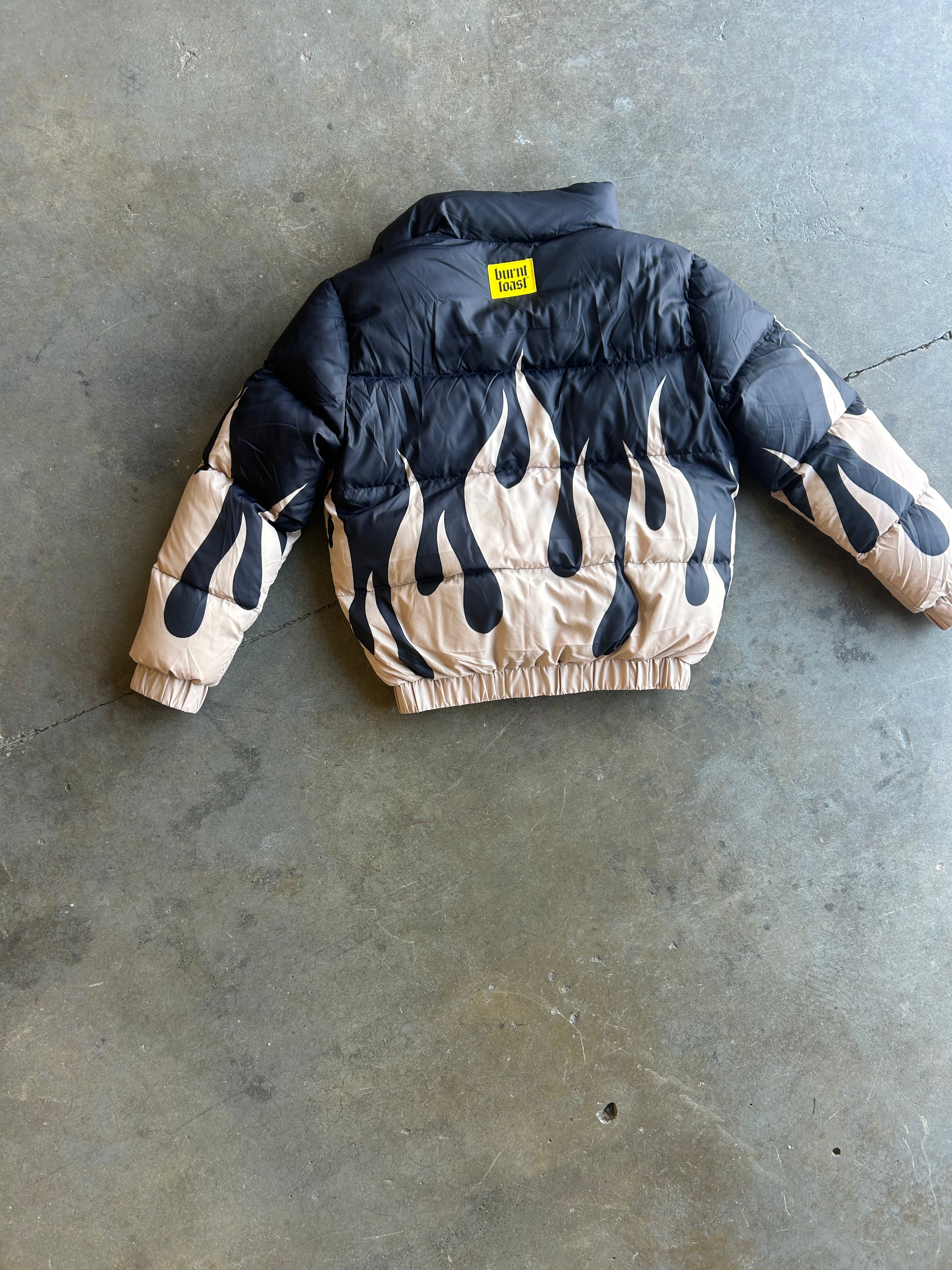 A puffer jacket laid flat on a concrete floor, featuring a black upper body and sleeves with a beige flame pattern on the lower body and cuffs. A small yellow label is visible at the back collar