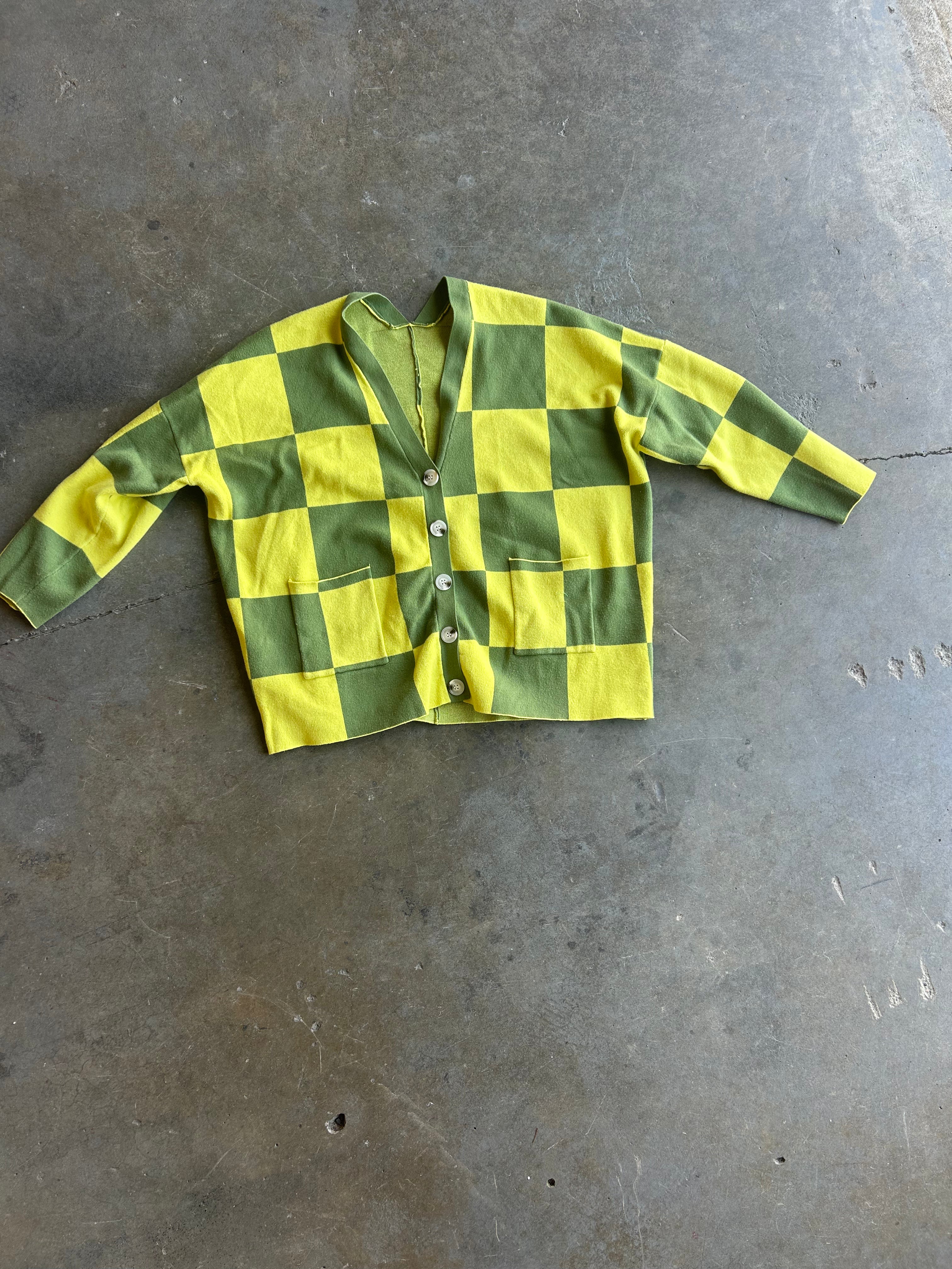 A yellow and green checkered cardigan sweater with a button-up front, laid flat on a gray concrete floor