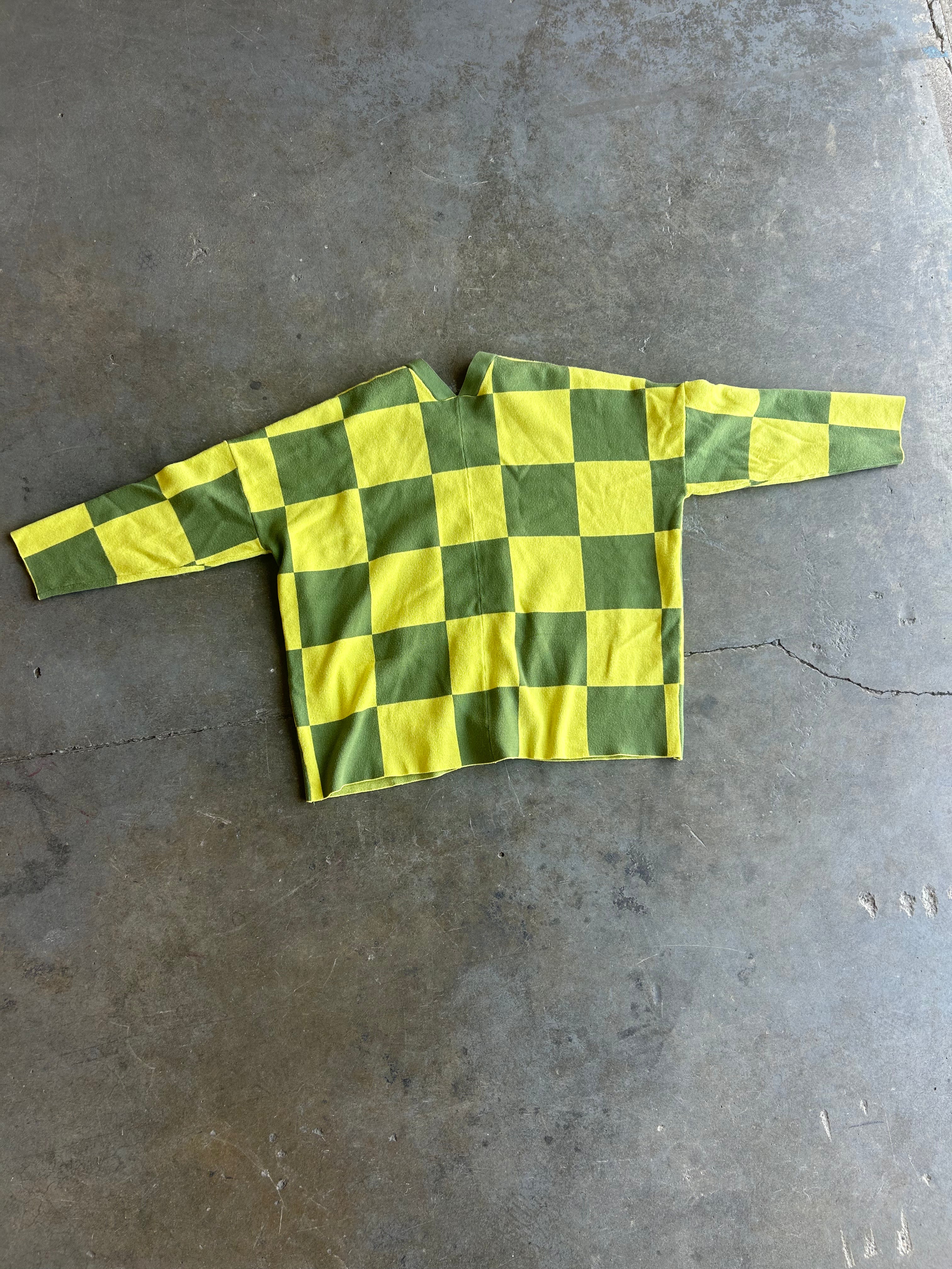 A long-sleeve shirt with a green and yellow checkerboard pattern, laid flat on a gray concrete floor