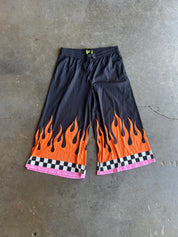 Black wide-leg shorts with a vibrant orange and yellow flame print at the hem, finished with a pink and black checkered border, laid flat on a gray concrete floor
