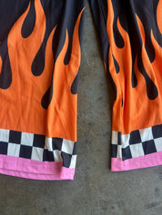 Close-up of flame-print shorts laid flat on a gray concrete surface. The shorts feature a bright orange and black flame pattern, a black and white checkered stripe, and a pink hem