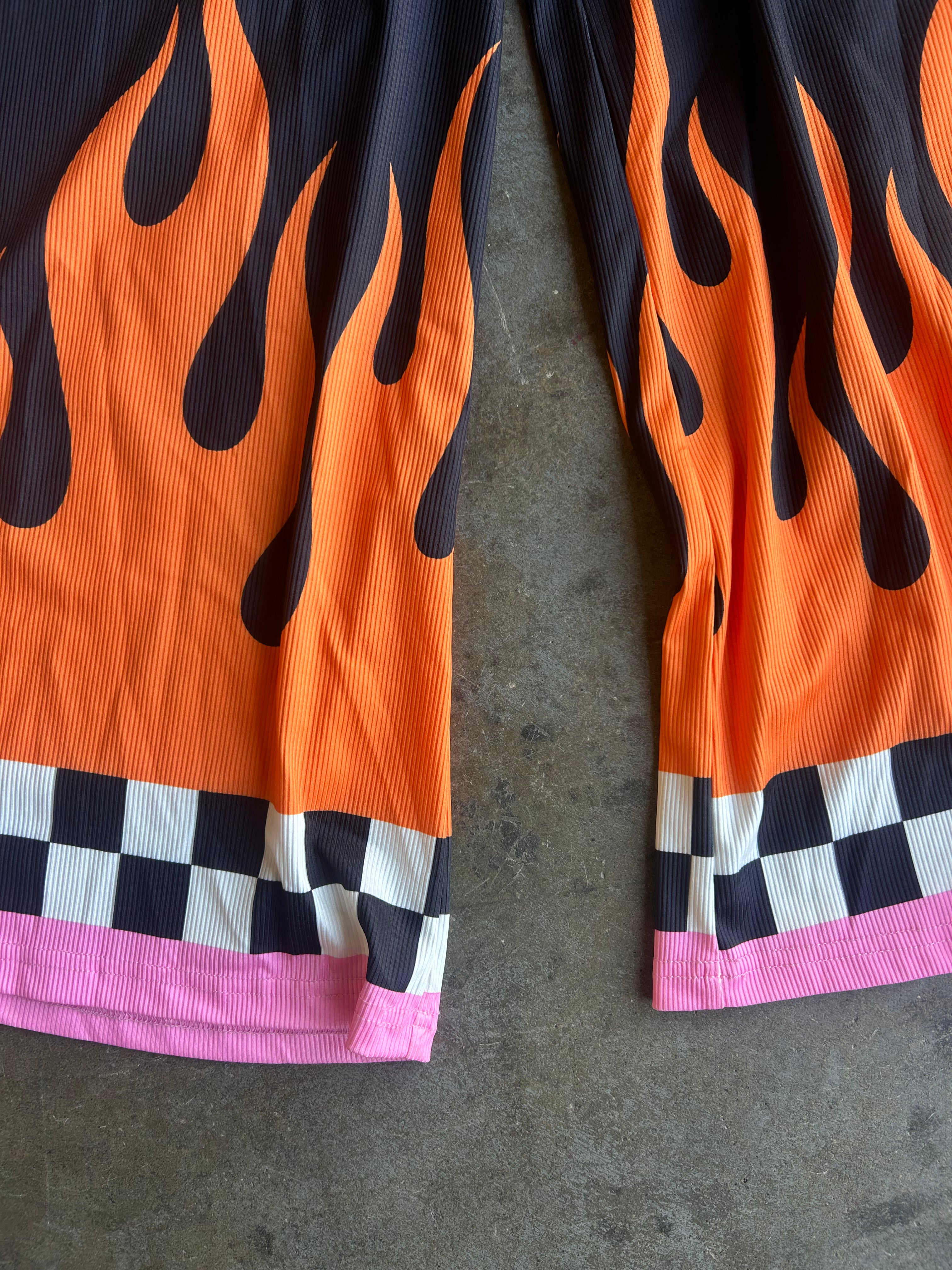 Close-up of flame-print shorts laid flat on a gray concrete surface. The shorts feature a bright orange and black flame pattern, a black and white checkered stripe, and a pink hem
