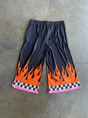 Black athletic shorts with a vibrant orange and black flame print near the hem, finished with a pink and black checkered border, laid flat on a gray concrete floor