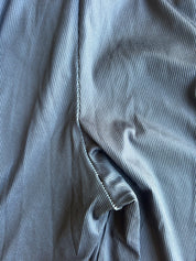 A close-up of draped slate blue corduroy fabric, showing the fine vertical ribbed texture and a neat white zigzag stitch along the edge