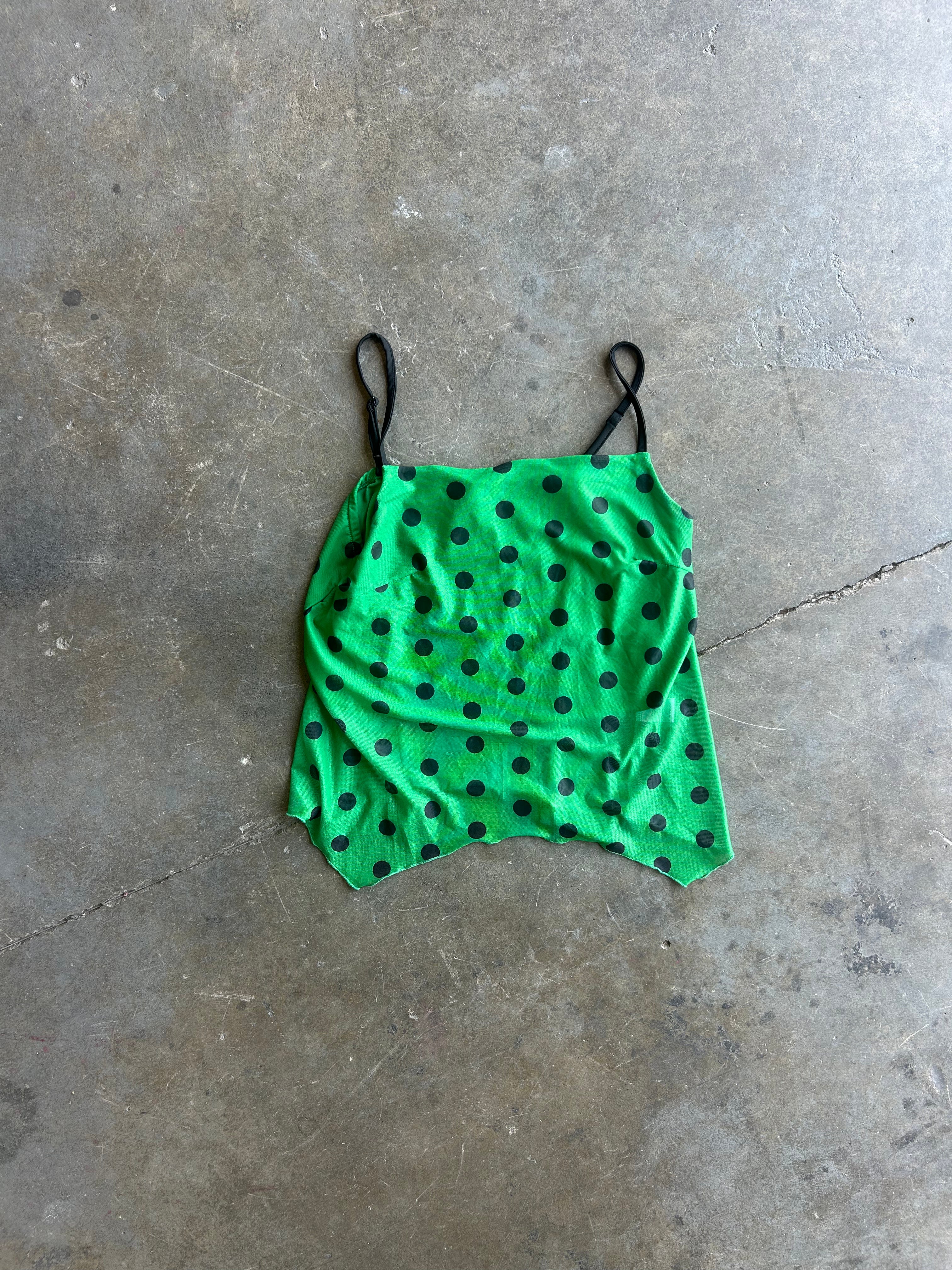 A bright green camisole top with black polka dots and thin black spaghetti straps, laid flat on a textured concrete floor