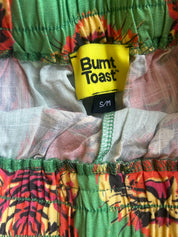Close-up of a garment's elastic waistband showing a yellow Burnt Toast brand label and a colorful tropical leaf print in green, orange, yellow, and red