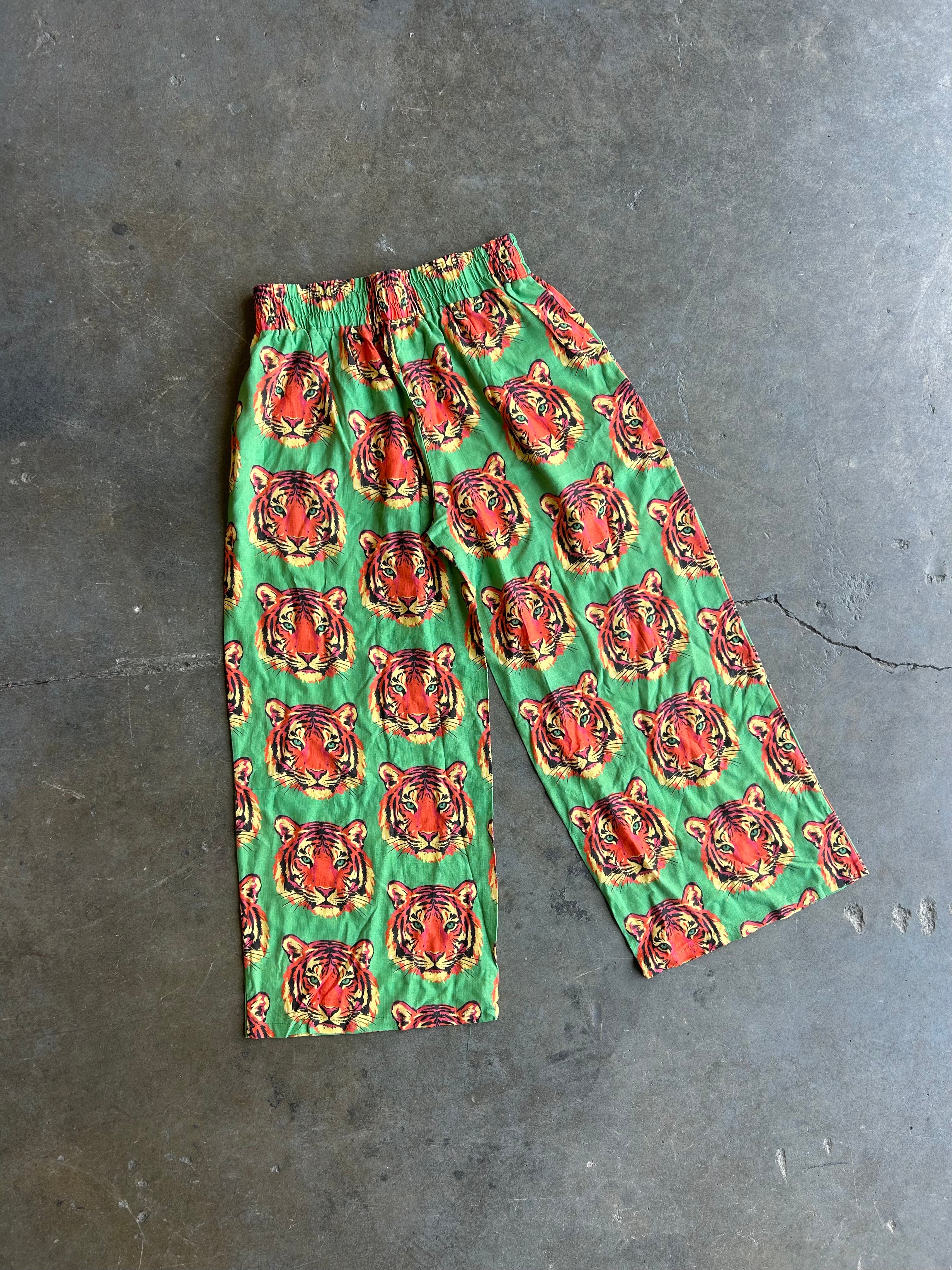 Green pants with a repeating red and orange tiger face pattern, laid flat on a gray concrete floor