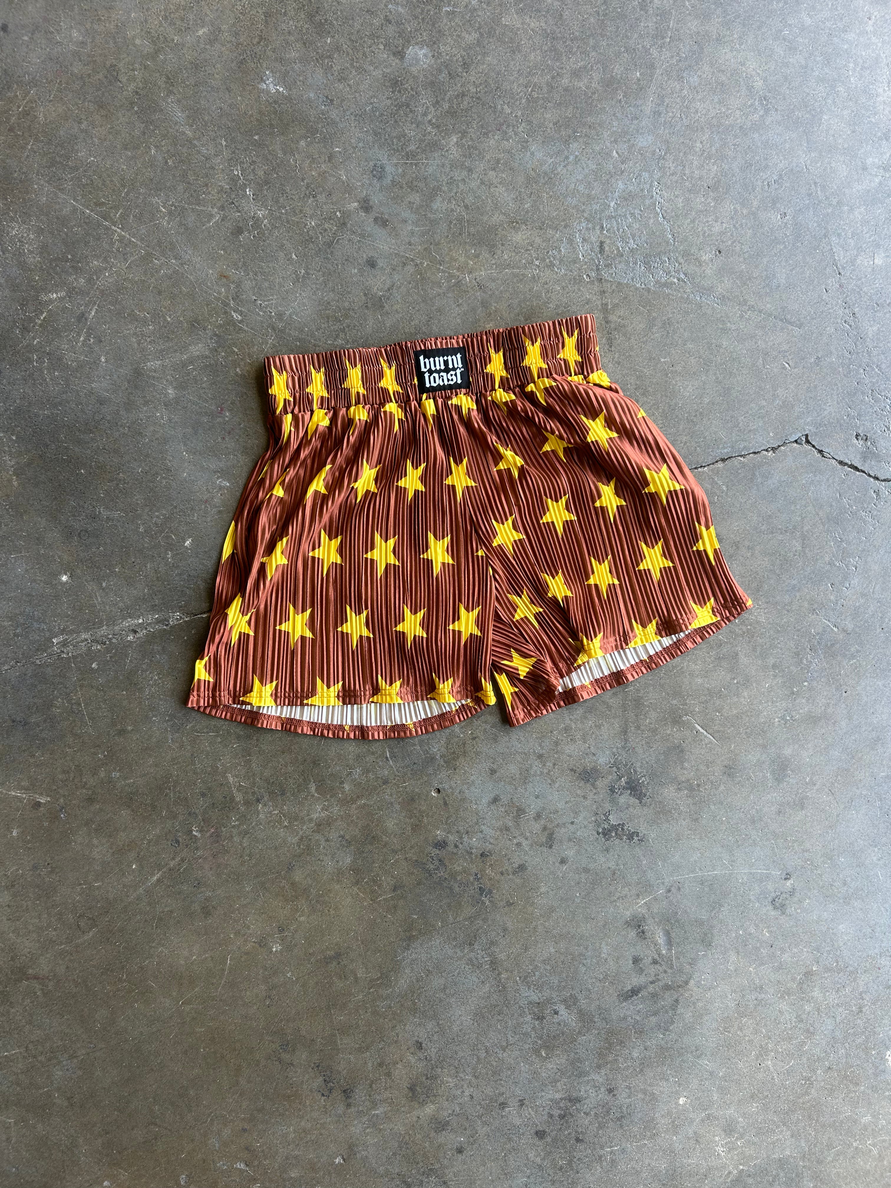 A pair of brown boxer briefs with a yellow star pattern, laid flat on a gray concrete floor. A small black brand label is visible at the waistband