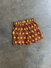 A pair of pleated shorts in rust brown with a yellow star pattern, laid flat on a gray concrete floor