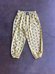 A pair of yellow jogger pants with an all-over black star pattern, laid flat on a dark textured surface. The pants feature an elastic waistband and elasticized cuffs
