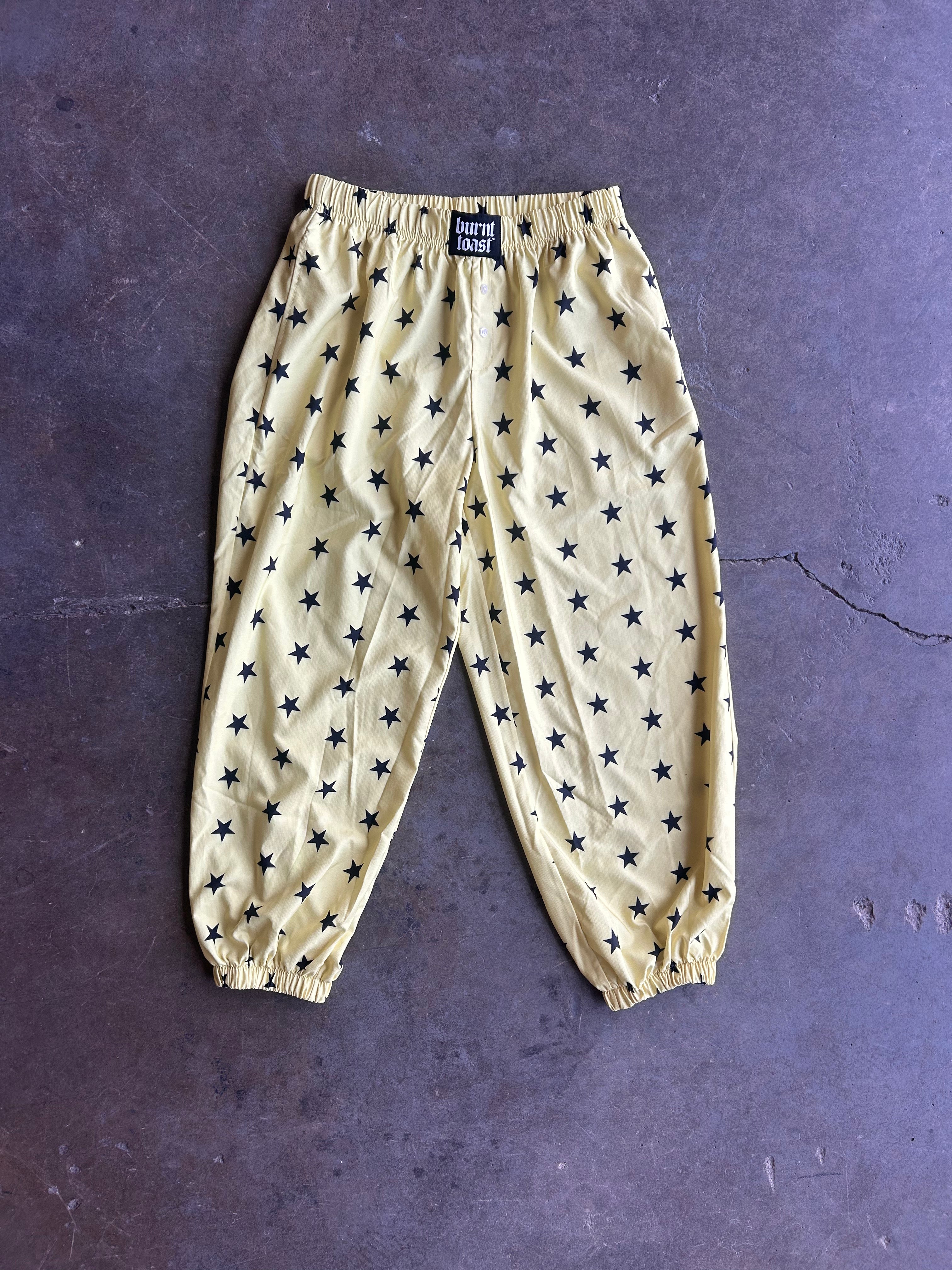 A pair of yellow jogger pants with an all-over black star pattern, laid flat on a dark textured surface. The pants feature an elastic waistband and elasticized cuffs