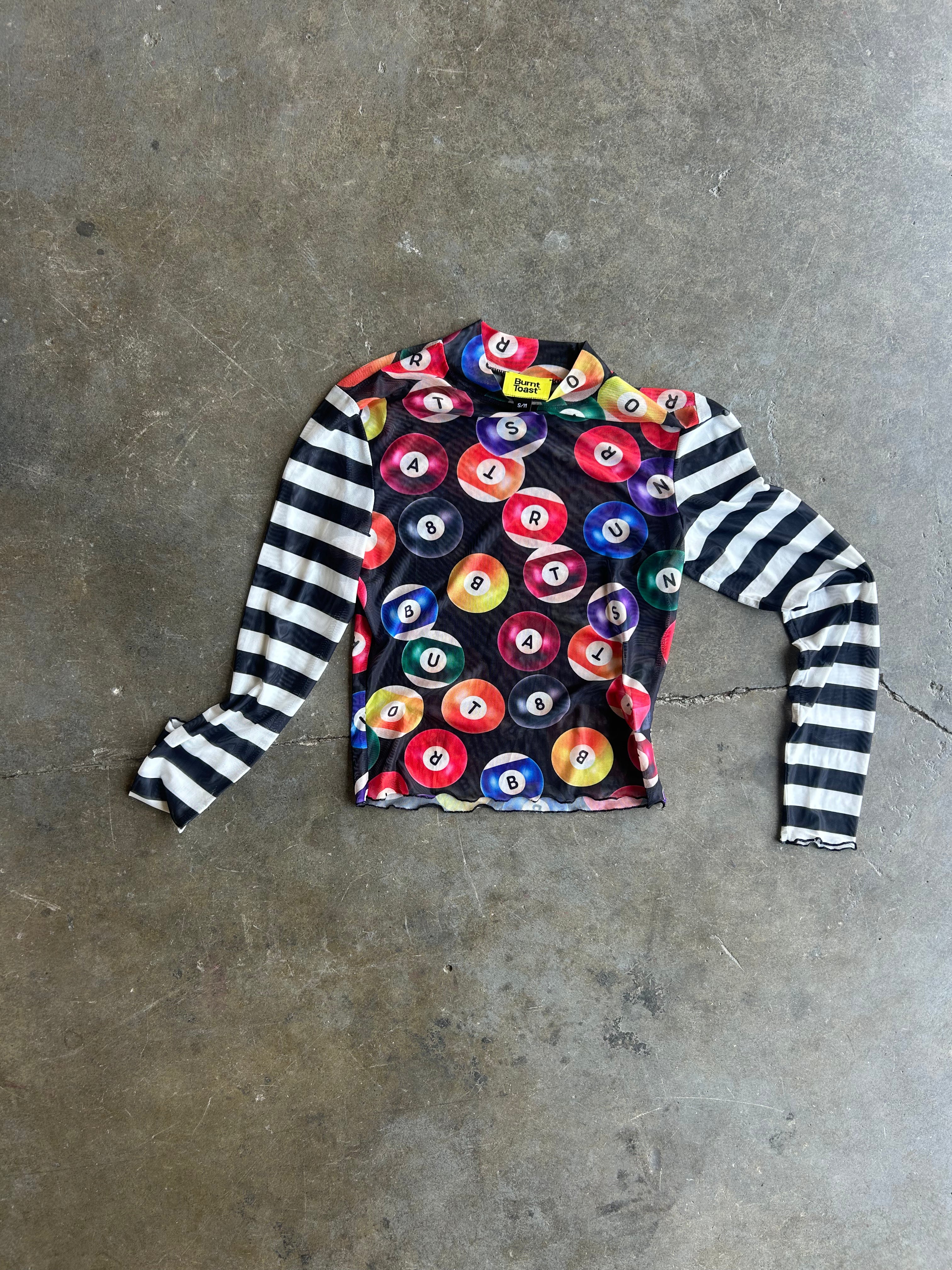 A long-sleeve shirt laid flat on a concrete floor, featuring a black body with a colorful pool ball pattern on the chest and black-and-white striped sleeves