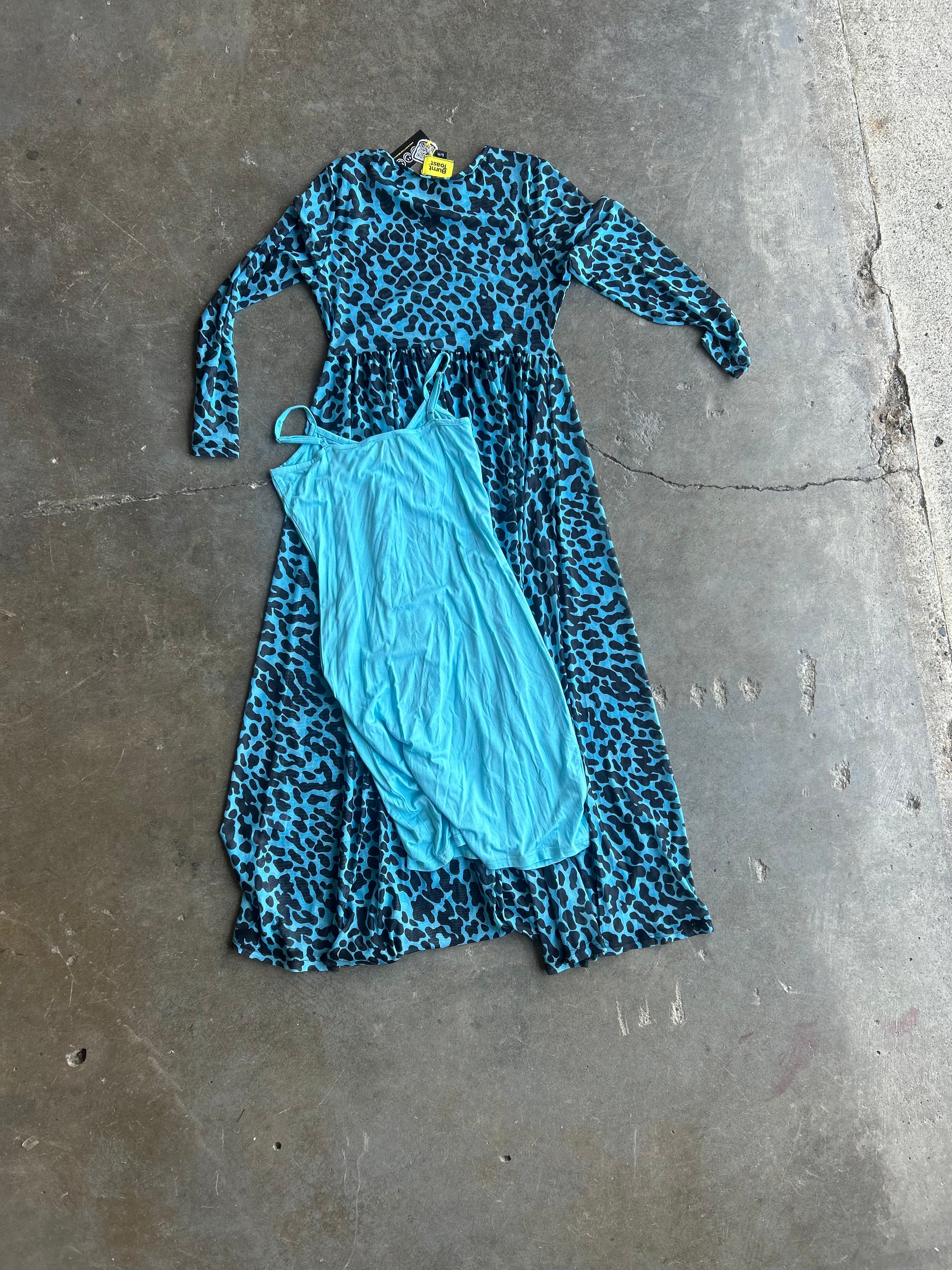 A long-sleeve maxi dress in a blue and black leopard print pattern, laid flat on a concrete floor next to a solid light blue slip dress