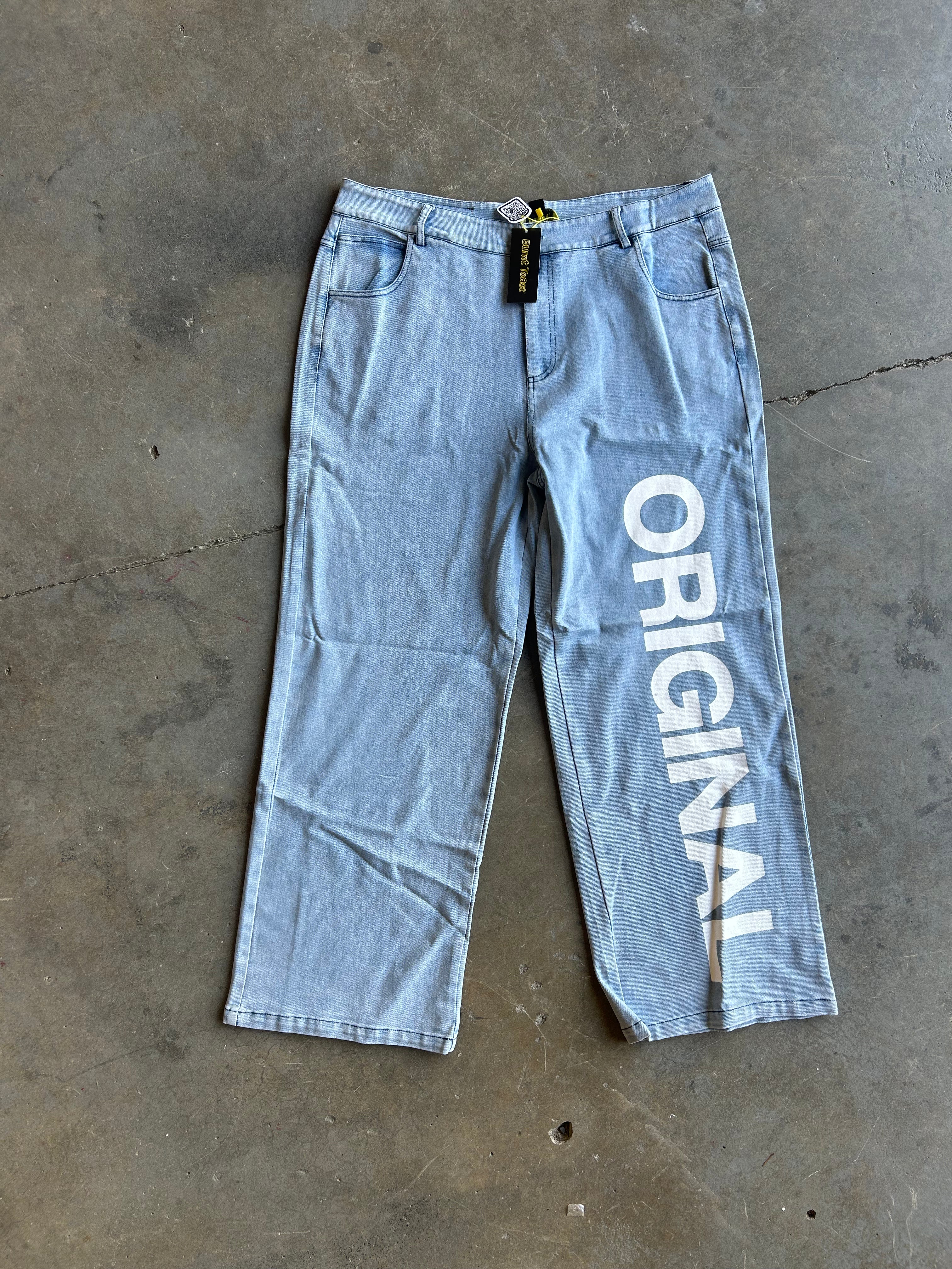 Light wash blue denim jeans laid flat on a concrete floor, featuring a large white vertical 'ORIGINAL' text graphic printed on the left leg