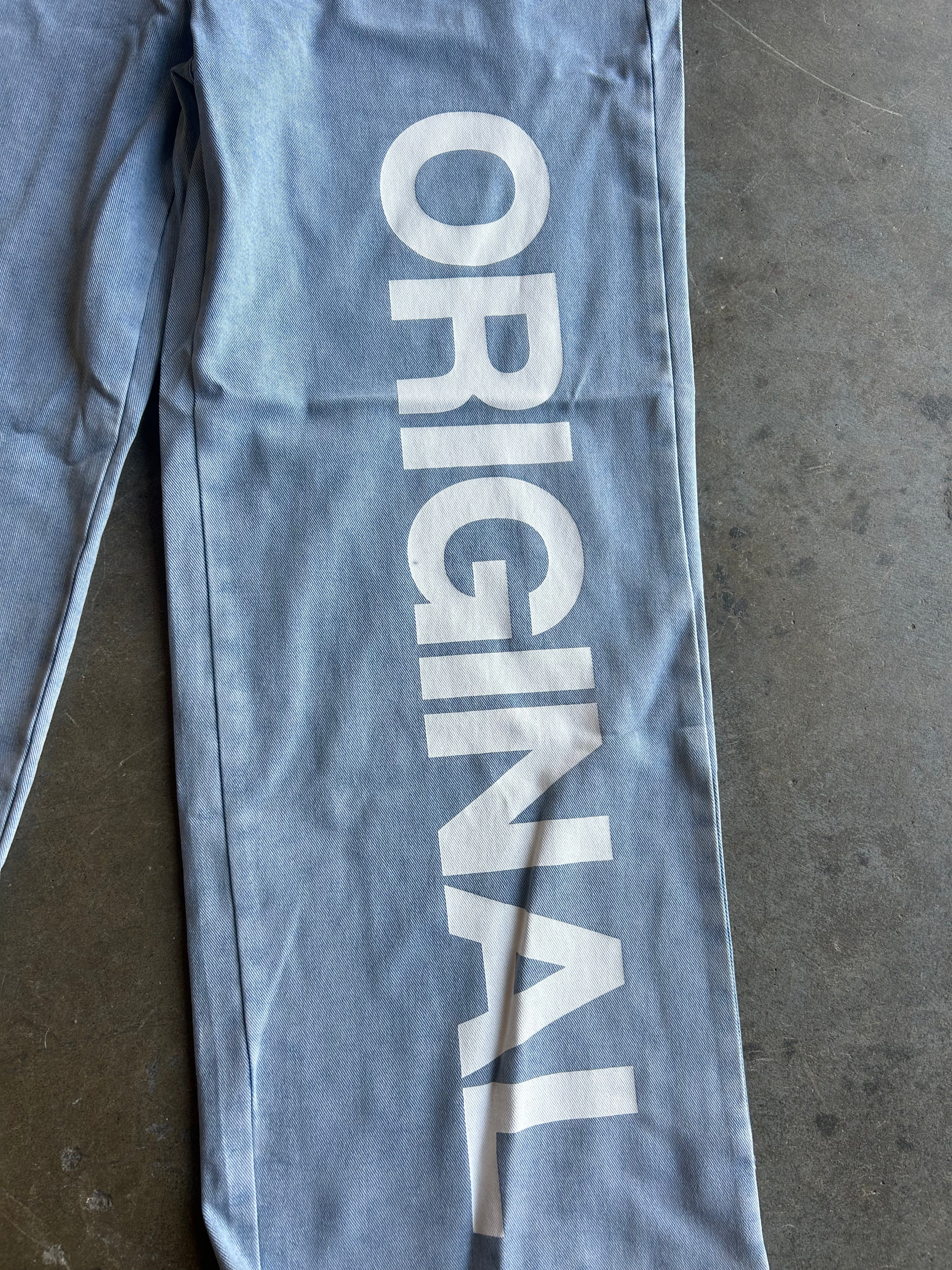 Light blue denim pants laid flat on a gray concrete floor, featuring the word 'ORIGINAL' printed vertically in large white letters down the left leg