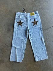 Light wash denim cropped jeans with star-shaped leopard-print patch back pockets, laid flat on a concrete floor
