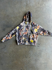 A camouflage zip-up hoodie with yellow stars and a yellow drawstring, laid flat on a gray concrete floor